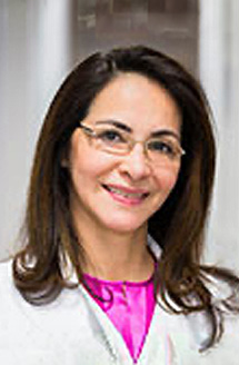 The image features a woman wearing glasses, smiling at the camera, with her hair pulled back. She is dressed professionally, wearing a white shirt and a pink top underneath, complemented by a light-colored blazer. Her attire suggests she may be in a professional setting or role.