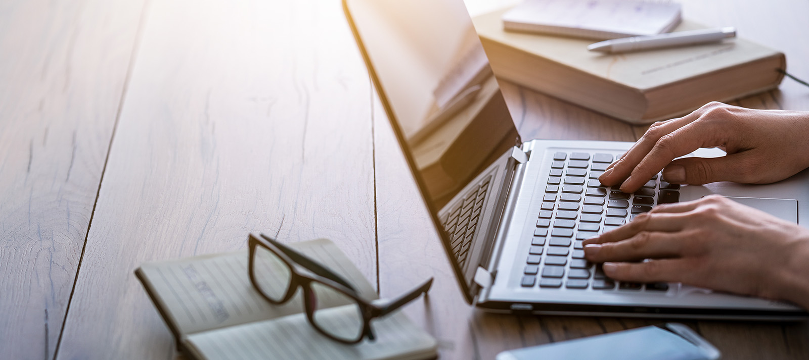 A person typing on a laptop computer with their hands on the keyboard, situated on a desk with various items around it, set against a blurred background.