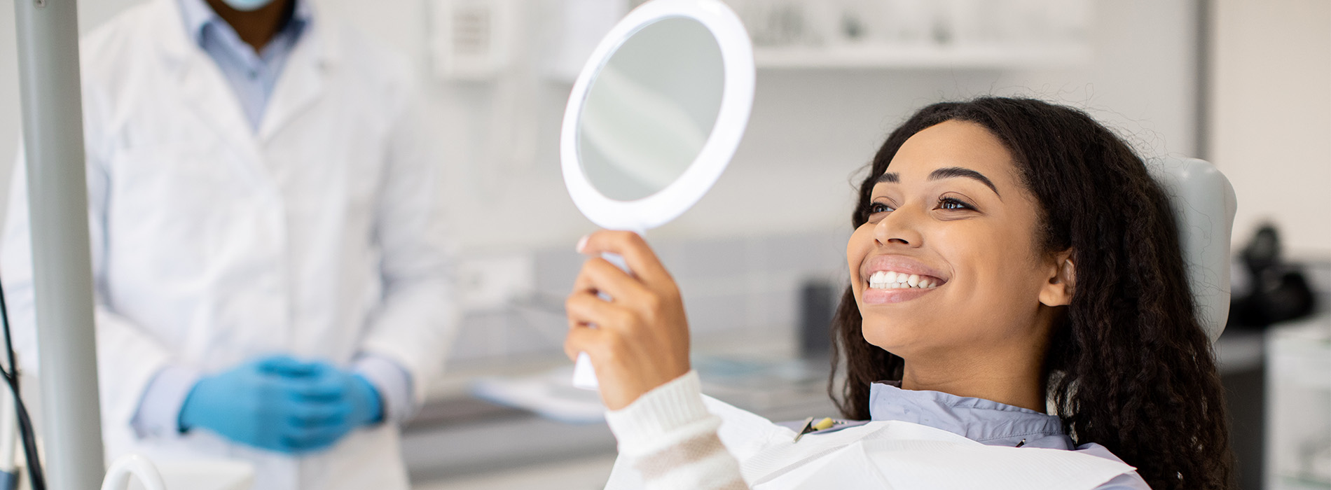 A woman with a bright smile sits in a dental chair, looking directly at the camera, while a dentist stands behind her, both in a professional dental office setting.