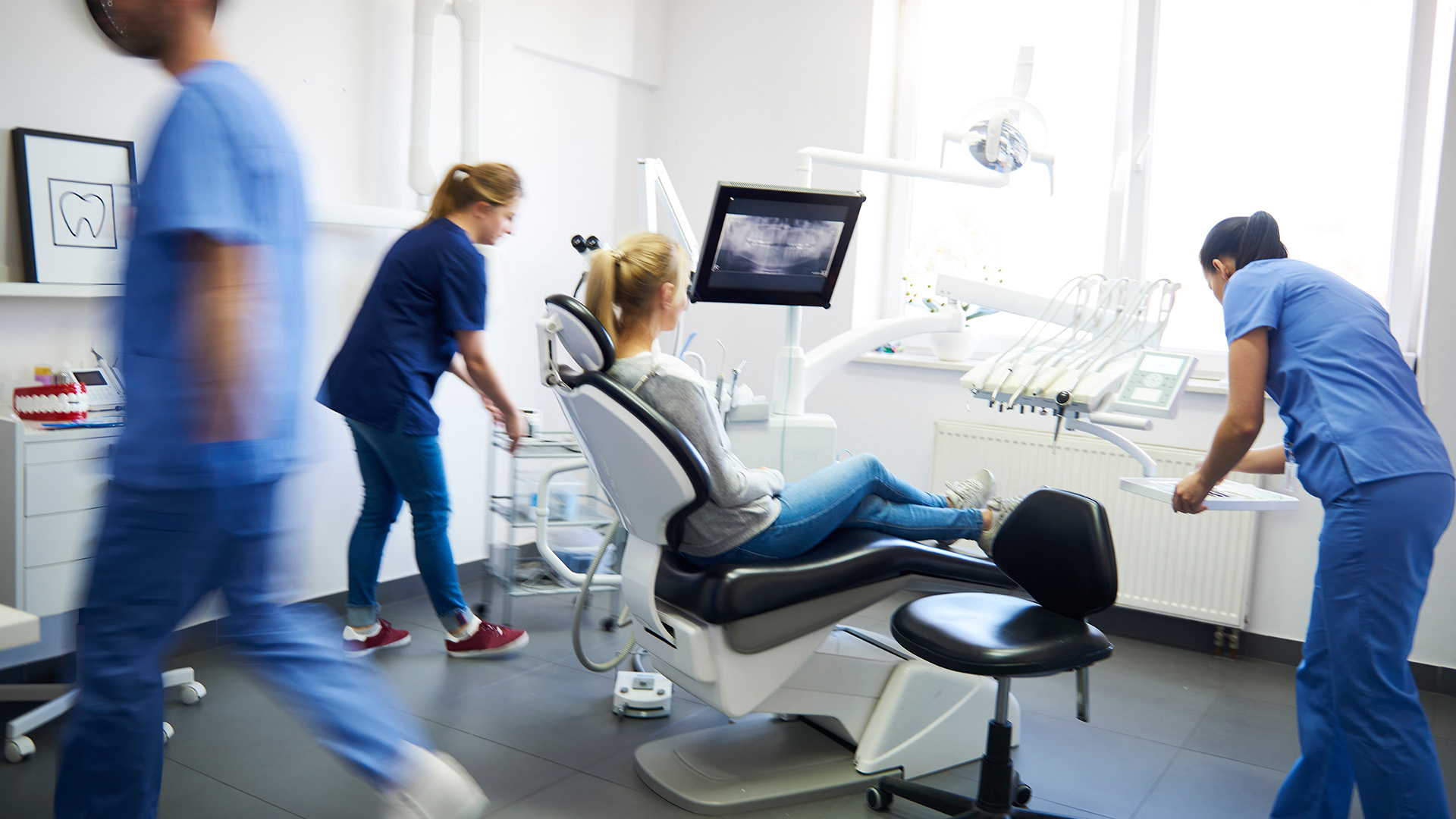 The image depicts a medical office with healthcare professionals attending to their duties, including a dentist s chair and dental equipment, and two individuals walking past a reception desk.