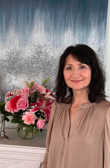 The image features a woman standing next to a table with flowers on it, posing for a photograph in front of a wall with a textured design.