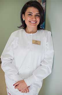 The image features a woman standing indoors with a smile, wearing a white lab coat and name tag, posing for a portrait.