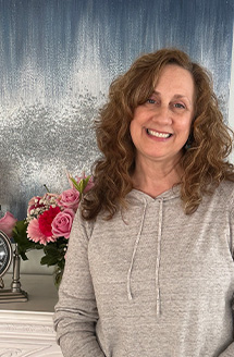 A smiling woman posing in front of a floral arrangement at a table with a clock on it.
