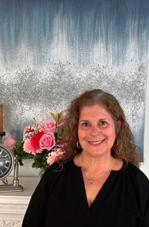 A woman standing in front of a wall with textured wallpaper, smiling at the camera, positioned next to a clock, flowers, and a candle.