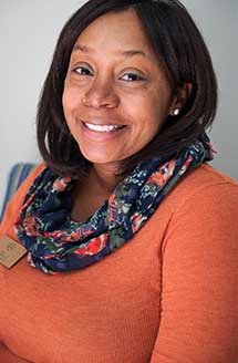 The image shows a smiling woman wearing an orange top with a black scarf around her neck, posing for a portrait with a neutral background.
