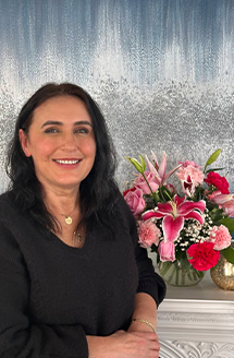 A smiling woman stands in front of a floral arrangement and a waterfall-style backdrop, posing for a portrait with a confident expression.