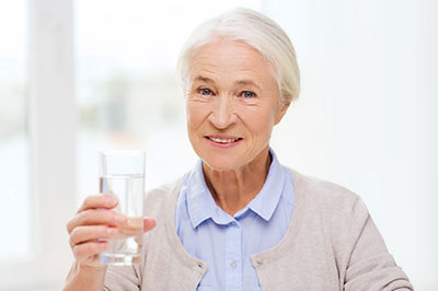 The image shows a woman holding a glass of water while standing indoors, smiling towards the camera.