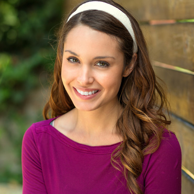 A smiling woman with long hair, wearing a purple top, standing against a wooden fence.