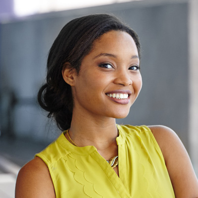 A smiling woman with dark hair wearing a yellow top and posing against a wall.