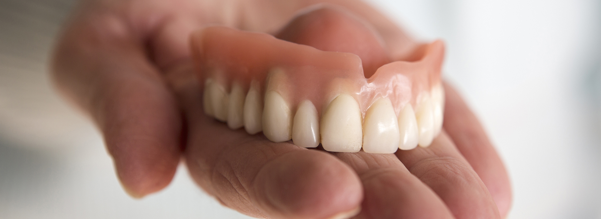 The image shows a person holding a set of dentures with visible teeth, against a blurred background.